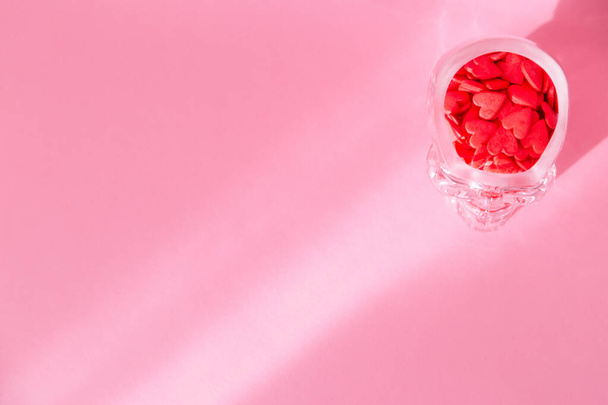 Una copa de vidrio en forma de cráneo humano está llena de corazones rojos y está sobre un fondo rosa
 - Foto, Imagen