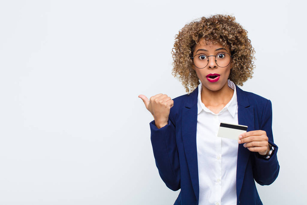 young african american woman looking astonished in disbelief, pointing at object on the side and saying wow, unbelievable with a credit card - Photo, Image