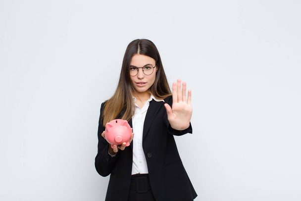 young pretty woman looking serious, stern, displeased and angry showing open palm making stop gesture with a piggy bank - Фото, изображение