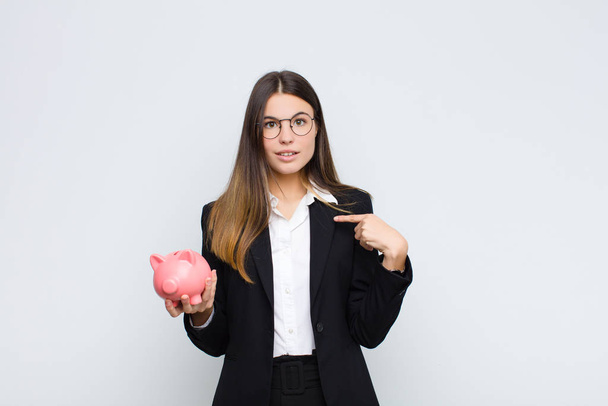 young pretty woman feeling happy, surprised and proud, pointing to self with an excited, amazed look with a piggy bank - 写真・画像