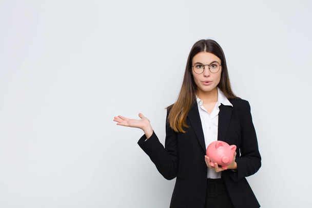 young pretty woman looking surprised and shocked, with jaw dropped holding an object with an open hand on the side with a piggy bank - Photo, Image