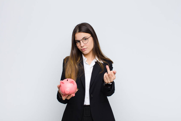 young pretty woman feeling angry, annoyed, rebellious and aggressive, flipping the middle finger, fighting back with a piggy bank - Photo, Image
