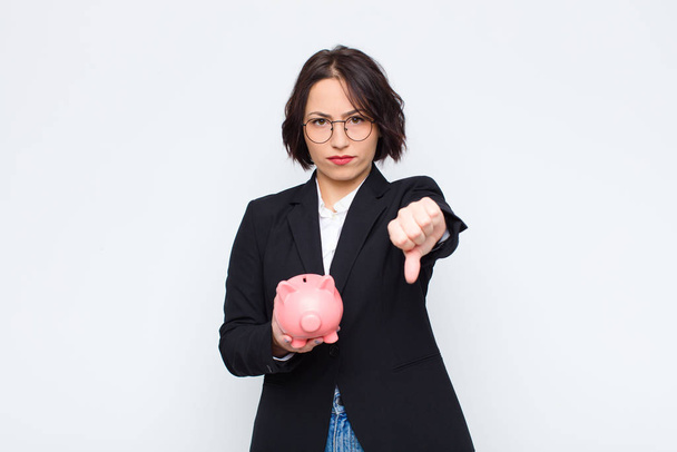 young pretty woman feeling cross, angry, annoyed, disappointed or displeased, showing thumbs down with a serious look with a piggy bank - Photo, image