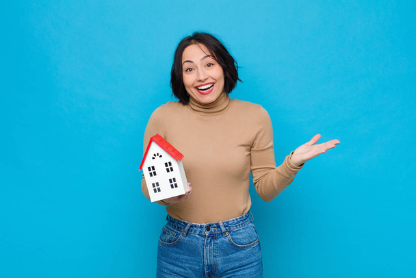 young pretty woman feeling happy, excited, surprised or shocked, smiling and astonished at something unbelievable with a house model - Fotoğraf, Görsel