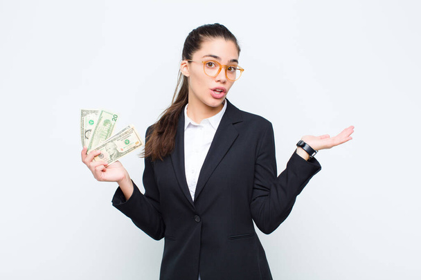 young businesswoman looking surprised and shocked, with jaw dropped holding an object with an open hand on the side with banknotes with bills - 写真・画像