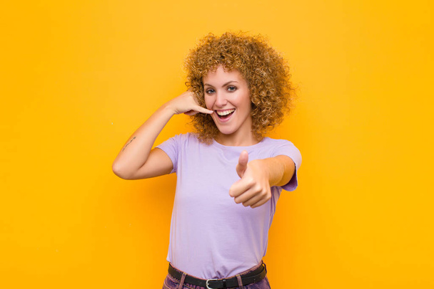 young afro woman smiling cheerfully and pointing to camera while making a call you later gesture, talking on phone against orange wall - Foto, Bild
