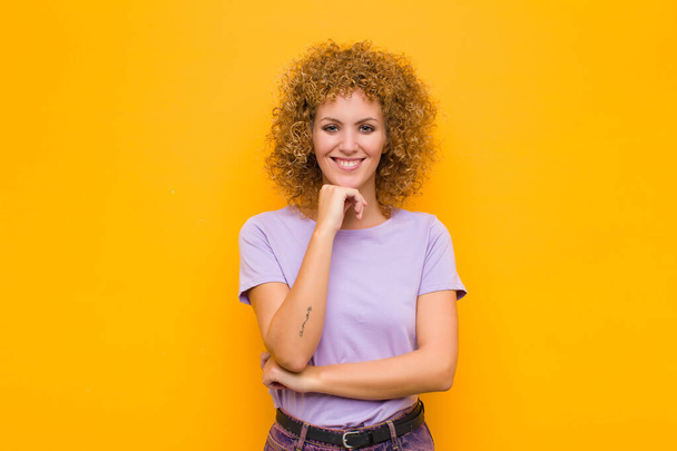 young afro woman looking happy and smiling with hand on chin, wondering or asking a question, comparing options against orange wall - Fotografie, Obrázek