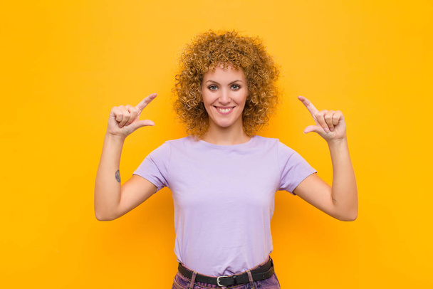 young afro woman framing or outlining own smile with both hands, looking positive and happy, wellness concept against orange wall - Photo, Image