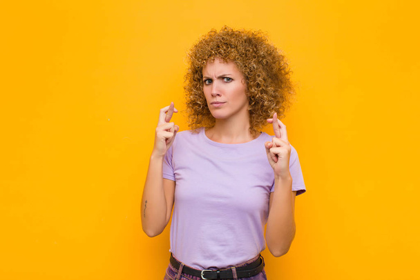 young afro woman crossing fingers anxiously and hoping for good luck with a worried look against orange wall - Фото, зображення