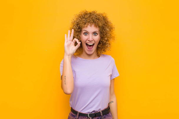 young afro woman feeling successful and satisfied, smiling with mouth wide open, making okay sign with hand against orange wall - Foto, Imagem