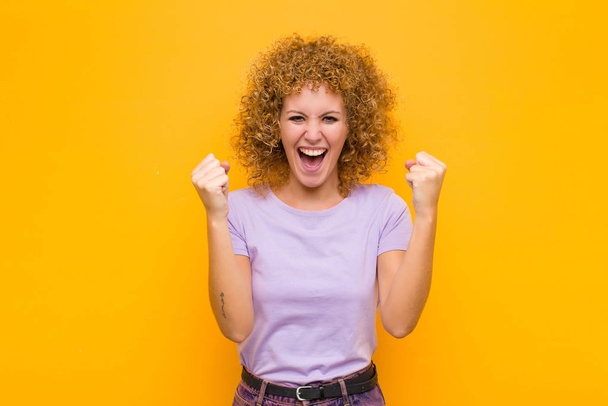 young afro woman feeling happy, surprised and proud, shouting and celebrating success with a big smile against orange wall - Fotoğraf, Görsel