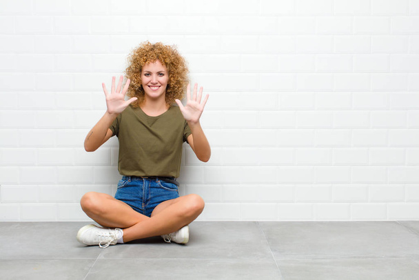 молодая афро женщина улыбается и выглядит дружелюбно, показывая число десять или десятую с рукой вперед, обратный отсчет, сидя на полу
 - Фото, изображение