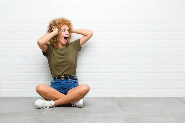 young afro woman with open mouth, looking horrified and shocked because of a terrible mistake, raising hands to head sitting on a floor - Fotografie, Obrázek