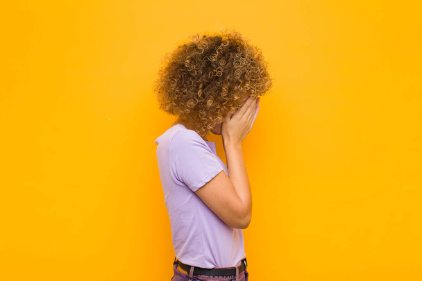 young afro woman covering eyes with hands with a sad, frustrated look of despair, crying, side view against orange wall - 写真・画像