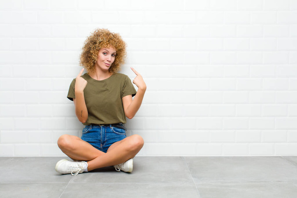 young afro woman with a bad attitude looking proud and aggressive, pointing upwards or making fun sign with hands sitting on a floor - Foto, Bild