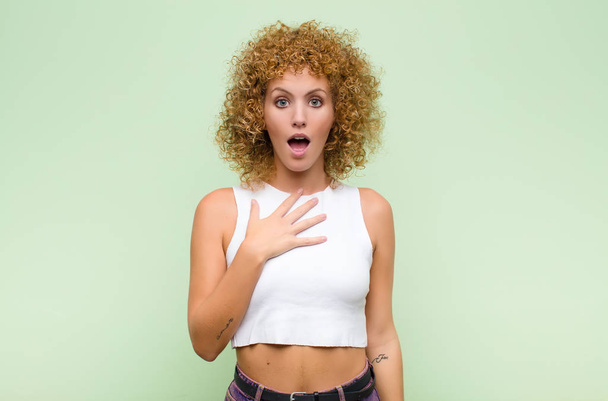 young afro woman feeling shocked, astonished and surprised, with hand on chest and open mouth, saying who, me? against green wall - Foto, Imagem
