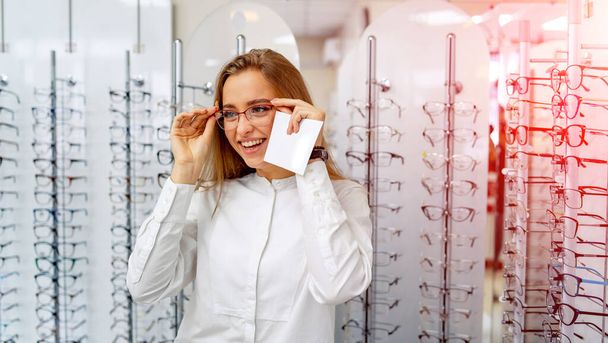 Happy female client or optician is standing with raw of glasses in background in optical shop. Stand with spectacles. Eyesight correction. Girl in glasses is posing to the camera. - Photo, image