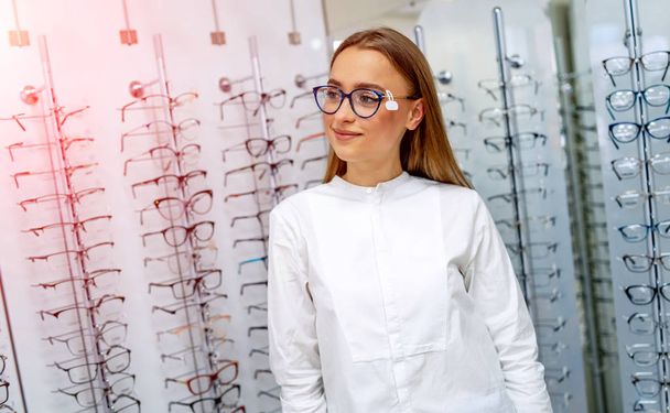 Model is standing with set of glasses in background in optical shop. Stand with spectacles. Eyesight correction. Girl wearing glasses. Portrait of a woman in correcting glasses. - 写真・画像