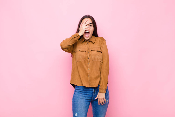 young pretty woman looking shocked, scared or terrified, covering face with hand and peeking between fingers against pink wall - Fotoğraf, Görsel