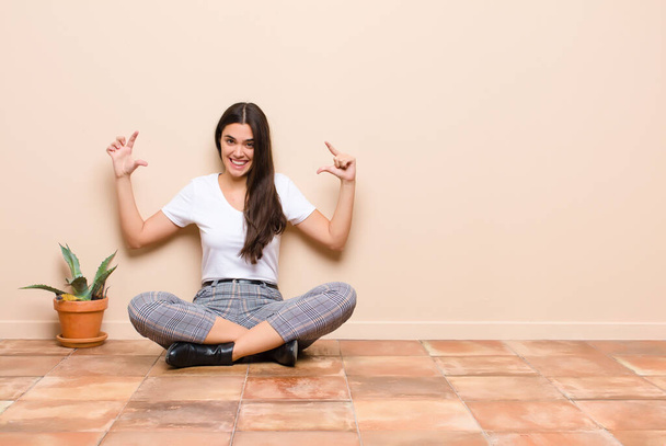 young pretty woman framing or outlining own smile with both hands, looking positive and happy, wellness concept sitting on a floor - Foto, imagen