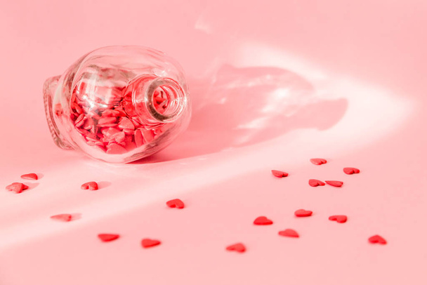 Red hearts pour out of a glass jug on a pink background - Foto, imagen
