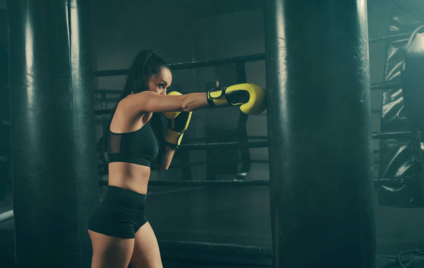 Ação tiro de jovem atlética mulher formação com saco de perfuração vestindo luvas de boxe amarelo
 - Foto, Imagem