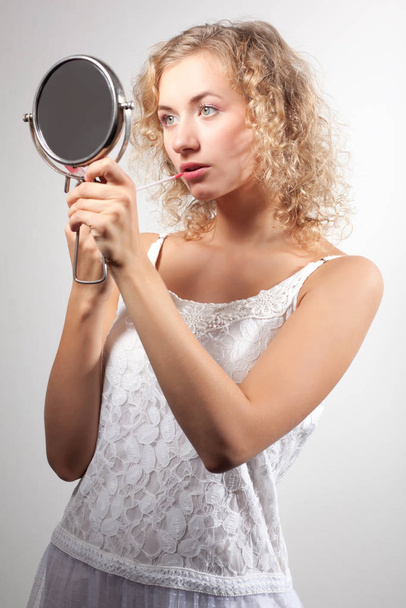 Young slim beautiful blond woman with curly hair in white sexy mini dress looking at small mirror and smiling - Foto, immagini