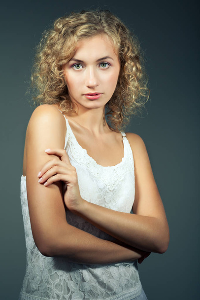 Young slim beautiful blond woman with curly hair in white sexy dress standing and looking at camera over grey background - Fotó, kép