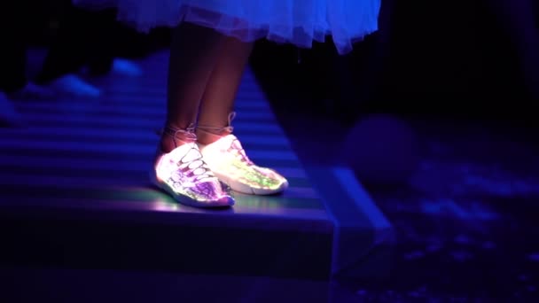 Le gambe di una ragazza in scarpe da ginnastica luminose bianche alla moda e gonna sul palco in un club scuro tra molte persone da vicino. La ragazza che rimane sulla scena e balla. La luce di scarpe illumina
. - Filmati, video