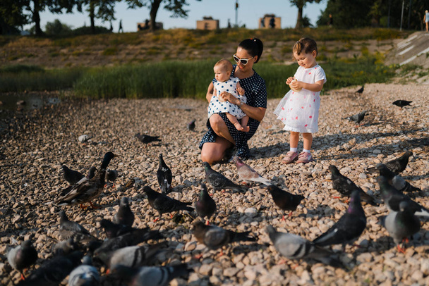 Jovem mãe com seu bebê menina filhas alimentando cisne e pequenos patinhos pássaros pão em um rio vestindo vestido pontilhado - Valores da família cor quente cena de verão
 - Foto, Imagem
