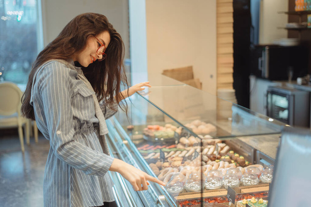 Krásná mladá Asiatka s dlouhými vlasy si vybere dezert v kavárně u okna. Krásný interiér pekařské kavárny - Fotografie, Obrázek