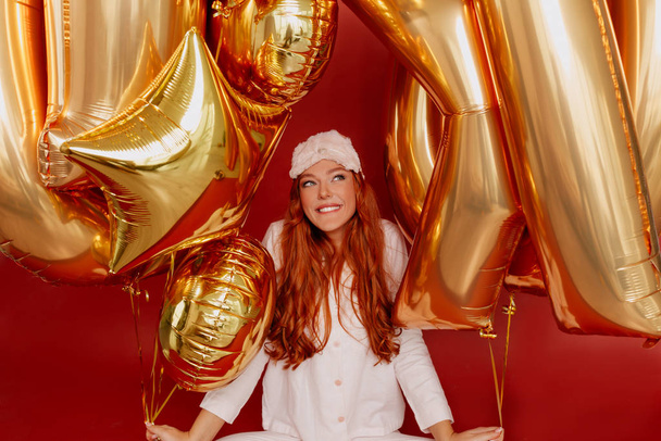 Retrato interior de una maravillosa dama de pelo rojo con pijamas rosas con globos dorados juguetones mirando hacia arriba y sonriendo durante la sesión de fotos sobre fondo rojo
 - Foto, Imagen
