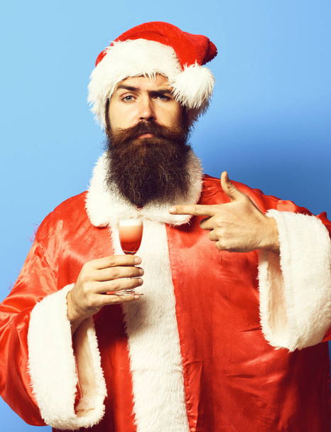handsome bearded santa claus man with long beard on serious face holding glass of alcoholic shot in christmas or xmas sweater and new year hat and shows on it on blue studio background - Foto, Imagem