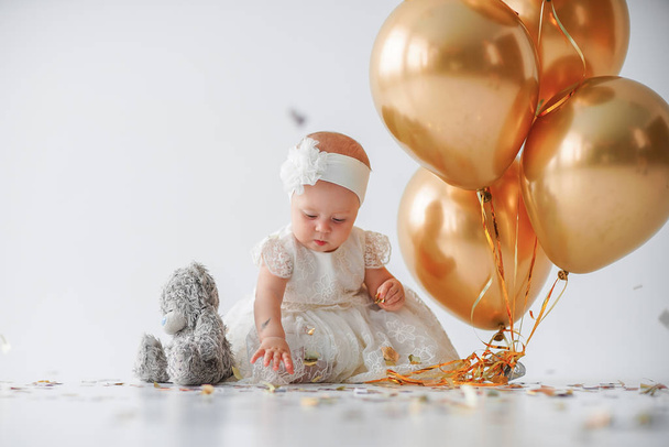 A one year old, baby girl sitting with a bunch of golden balloons and taddy bear on white background in studio - Fotografie, Obrázek