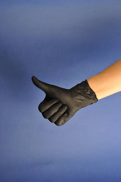 Hands in black latex gloves.  Thumb up, the remaining fingers clenched.  Blue background.  Close-up.  Concept: finger gestures. - Фото, изображение