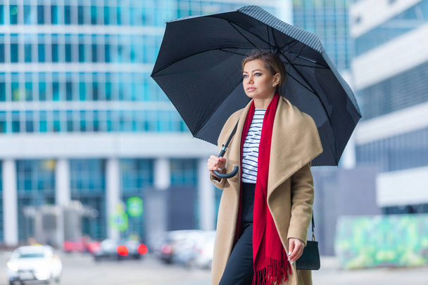 Rain. Outdoors lifestyle fashion portrait of stunning brunette girl. Walking on the city street. Going shopping. Wearing stylish white fitted coat, red neckscarf, black umbrella cane. Business woman. - Foto, afbeelding