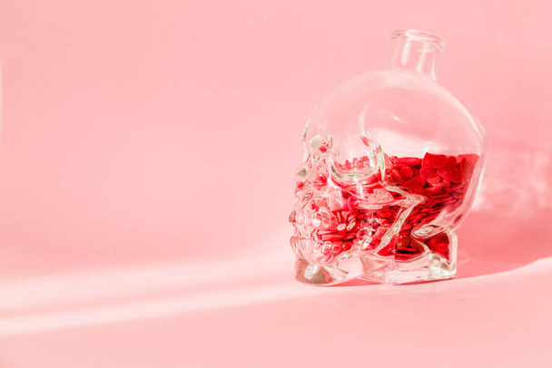 A glass carafe in the shape of a human skull is filled with red hearts and stands on a pink background - Photo, Image