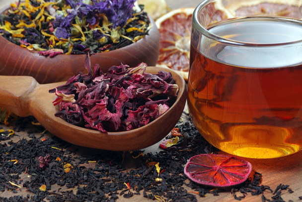 tazza di tè fresco e diverse varietà di foglie di tè su un tavolo di legno. tè miscelato con petali di fiori, ibisco in un cucchiaio di legno e agrumi secchi
.  - Foto, immagini