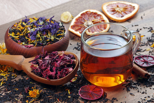 kopje verse thee en verschillende soorten theebladeren op een houten tafel. gemengde thee met bloemblaadjes, hibiscus in een houten lepel en gedroogde citrusvruchten.  - Foto, afbeelding