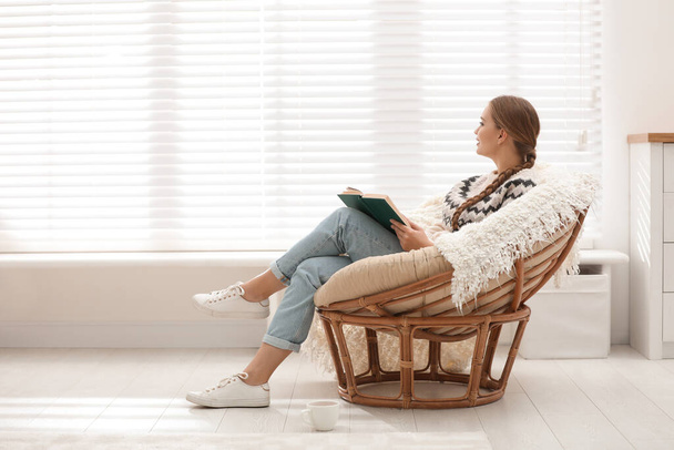 Young woman reading book in papasan chair near window at home - Valokuva, kuva