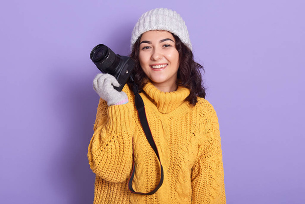 Portrait of beautiful attractive brunette holding photocamera in one hand, looking directly at camera, wearing yellow sweater, grey gloves and hat, having pleasant facial expression. Hobby concept. - Fotoğraf, Görsel