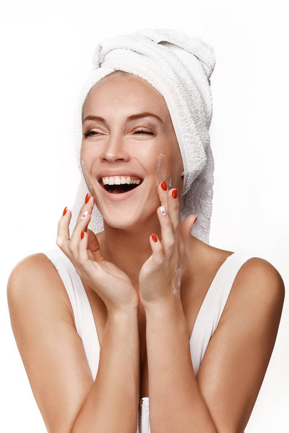 A beautiful young girl in a white blouse with a towel on her head, after a shower, applies cream to her face and laughs. Moistens a face with cream. Spa. In the studio on a white background. - Photo, Image