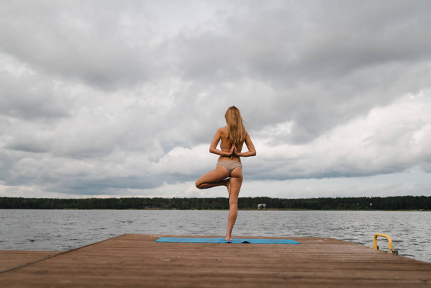 Тонкая женщина, практикующая йогу в бикини на озере с облаками - Yoga meditation and wellness lifestyle concept - European Eastern Latvia Riga
 - Фото, изображение