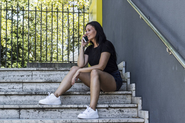 Женщина говорит по смартфону, сидя на лестнице, в городском парке
 - Фото, изображение