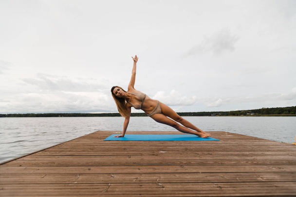 fitte schlanke Frau praktiziert Yoga-Übungen im Mini-Badeanzug-Bikini am wolkenverhangenen See - Yoga-Meditation und Wellness-Lifestylekonzept - Europäisches Ostlettland riga - Foto, Bild