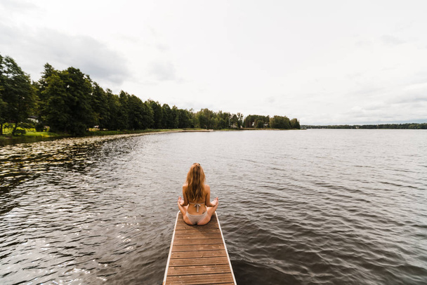 Fitt karcsú nő gyakorló jóga gyakorlatok visel mini fürdőruha bikini a tó felhők - jóga meditáció és wellness életmód koncepció - Full sitting shot - Fotó, kép