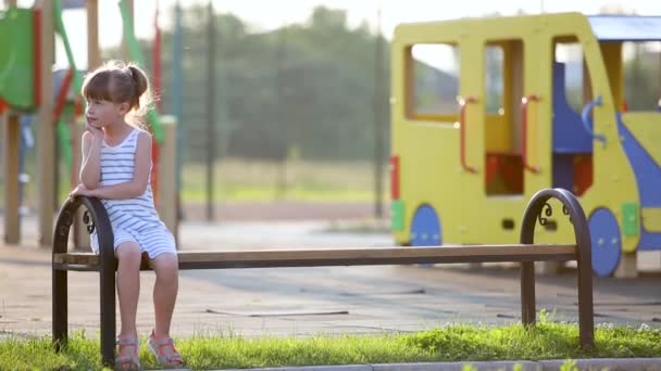 Nettes Mädchen, das auf einer Bank auf dem Sommerspielplatz im Kindergartenhof auf seine Mutter wartet. - Filmmaterial, Video