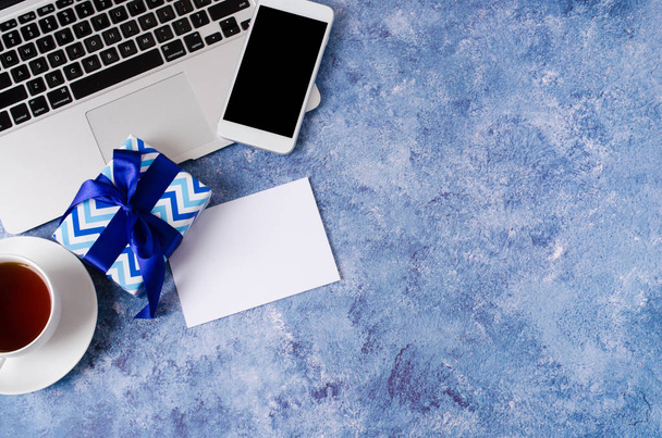 Blue gift box, smartfon z czarnym białym ekranem na biurku, laptop i filiżanka herbaty na niebieskim tle. - Zdjęcie, obraz