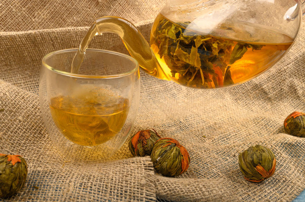 Τσάι λουλούδι παρασκευάζεται σε ένα ποτήρι τσαγιέρα, ένα ποτήρι τσάι και μπάλες τσαγιού λουλούδι σε ένα φόντο από τραχύ ύφασμα homespun. Κλείσε.. - Φωτογραφία, εικόνα