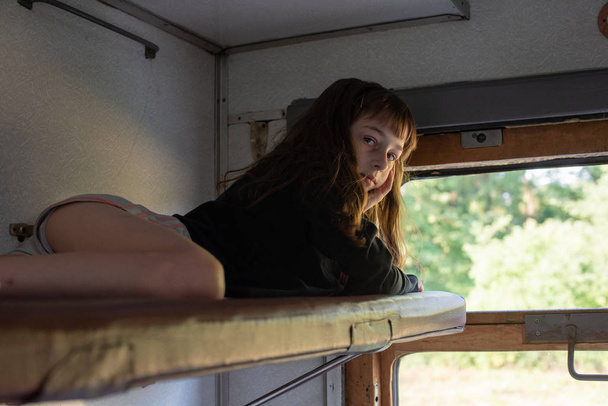Χαριτωμένο κορίτσι ξαπλωμένο στο πάνω ράφι του τρένου και κοιτάζοντας έξω από το παράθυρο στο δρόμο. Καλοκαιρινές διακοπές - Φωτογραφία, εικόνα